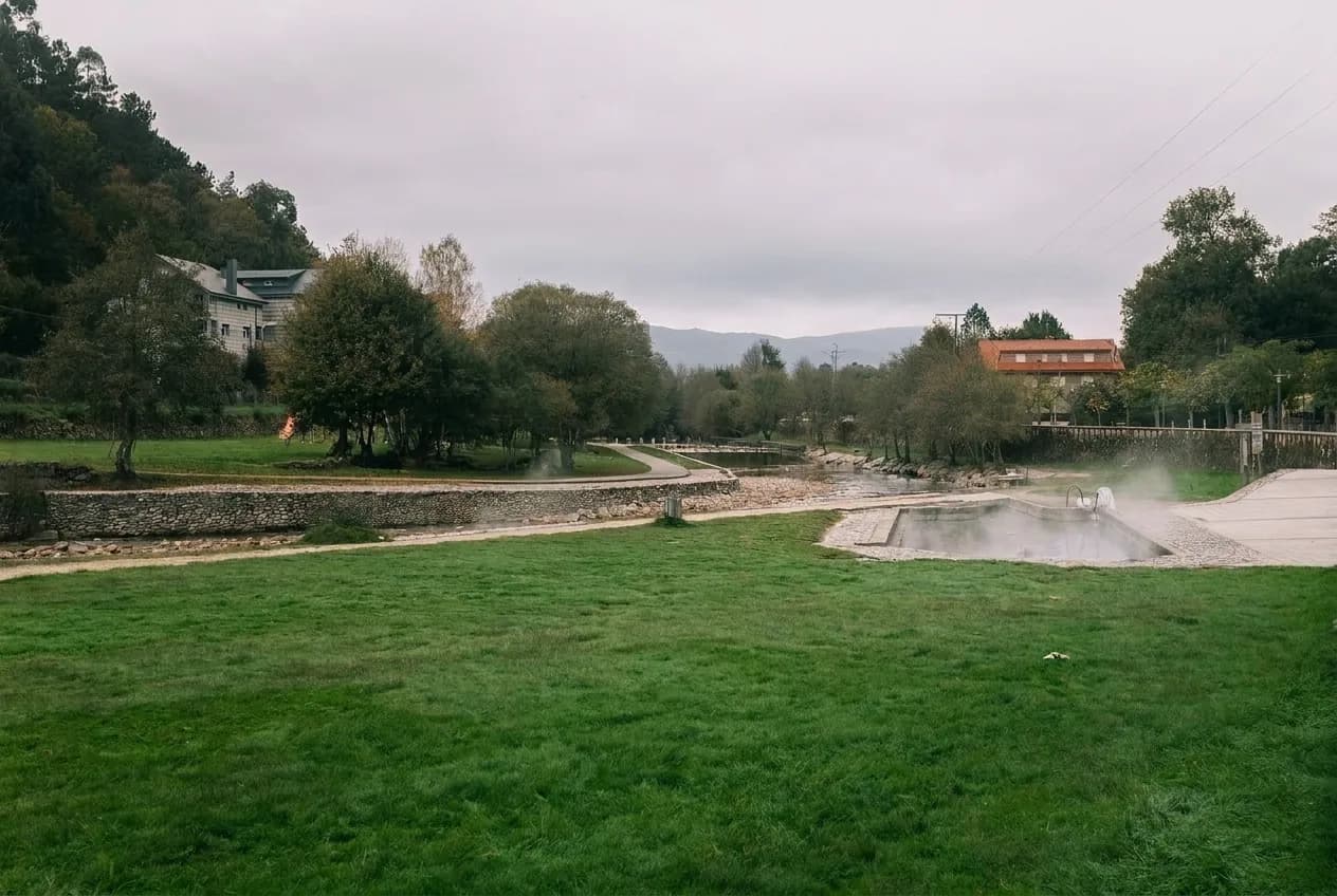 Pozas de Río Caldo en Os Baños con vapor sobre el agua, un prado delante y casas al fondo
